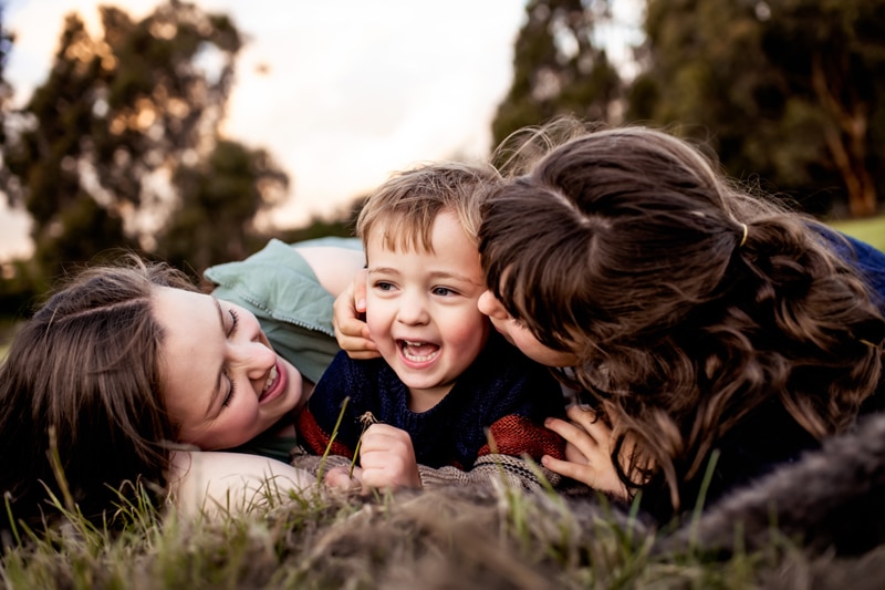 Family Photographer, two older sisters admire and play with their younger brother in the grass